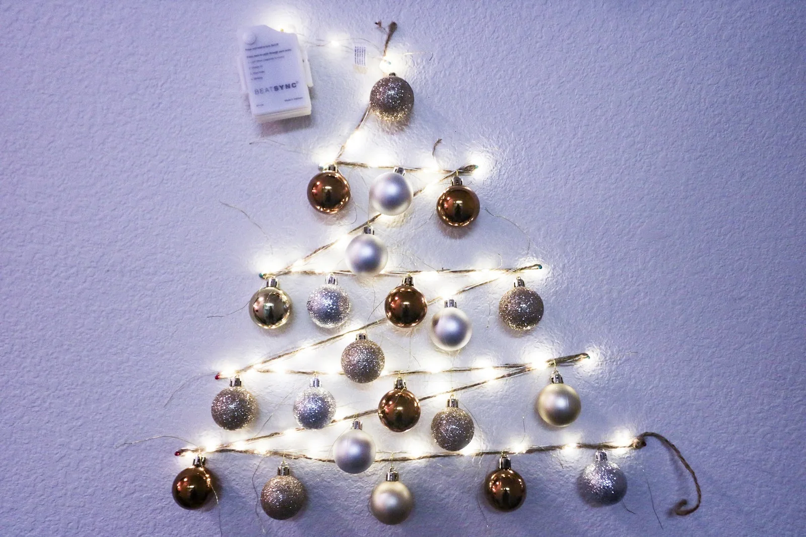 A Christmas tree outline made of fairy lights and hanging baubles on a bare wall, no nails used