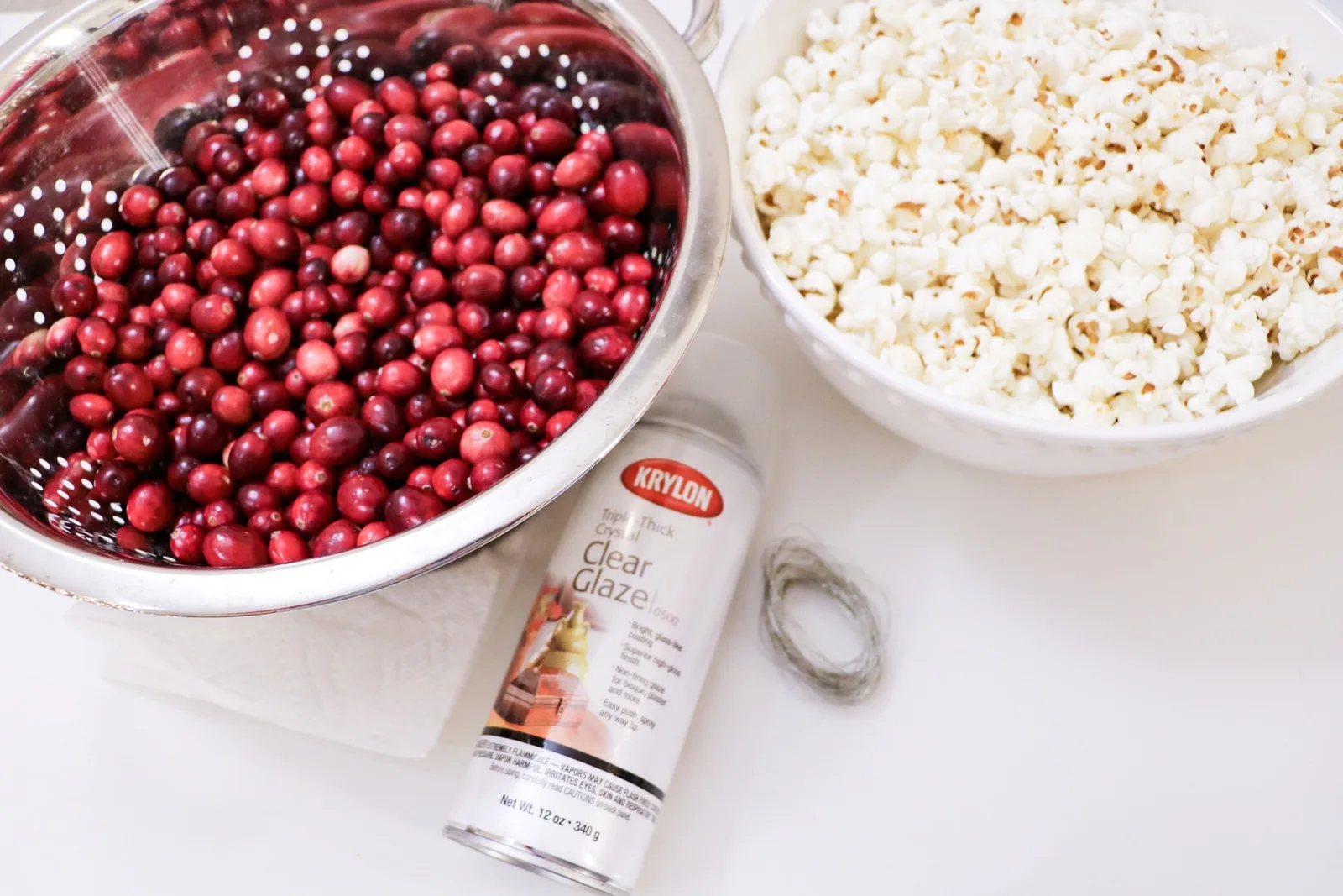 A colander of fresh cranberries, a bowl of air-popped popcorn, a can of Krylon clear glaze spray and a coil of floral wire on a white counter